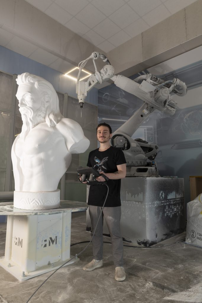 Operatore in laboratorio mentre controlla un braccio robotico per la fresatura di una grande scultura in polistirolo raffigurante un busto maschile.